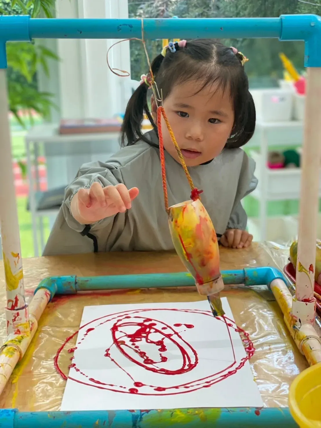 A toddler watching a swinging bottle of paint draw red circles on paper