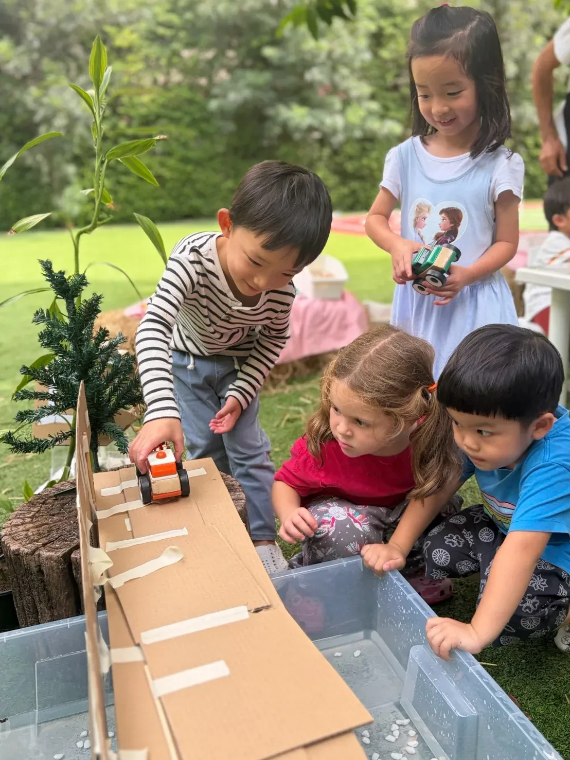 Four children watching a toy car run down the cardboard ramp they built