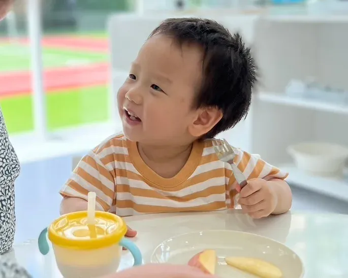 A smiling toddler eating fruit at snack time, with a teacher close by